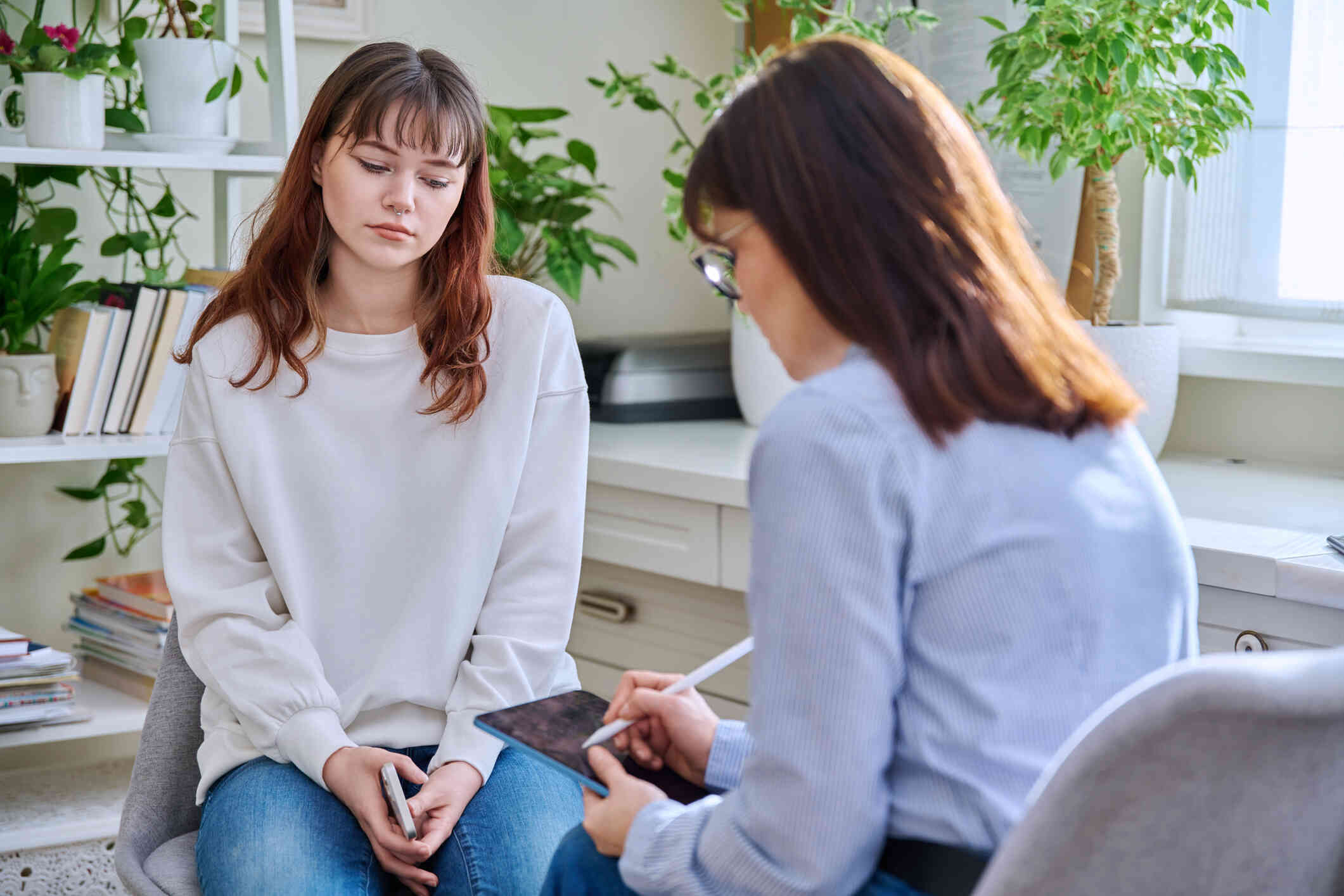 A woman in a white sweater sits sadly in a chair across from her female therapist as she writes on a tablet.